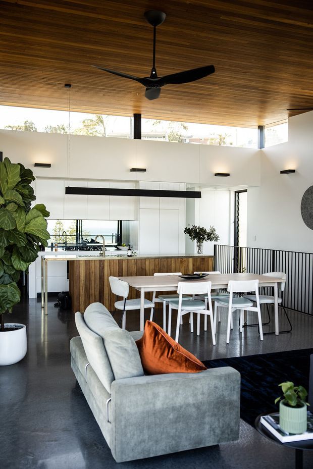 Open-plan contemporary living and dining area with a visible kitchen behind it. The room features a richly timber-lined ceiling, polished dark floor, white walls, a large black ceiling fan, linear pendant lighting, a timber kitchen island, white dining furniture, a pale grey sofa, and black metal balustrading around a stair opening. High clerestory-style glazing brings in daylight.