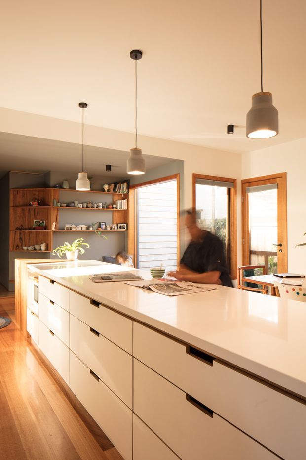 A bright, warm contemporary kitchen centered on a large white island with extensive integrated drawers and dark recessed pulls. Timber flooring, white cabinetry, and a polished light benchtop contrast with open timber shelving filled with kitchenware along the rear wall. Pendant lights hang from a smooth white ceiling, while timber-framed glazed doors and windows bring in natural light. A person is seated at the island, with newspapers and small objects on the benchtop.