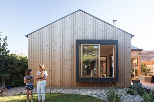 Front exterior view of a compact gable-roofed dwelling or extension clad in vertical pale timber boards. A large dark-framed window reveals part of the interior, while a simple landscaped garden with gravel and planting sits in front. The image has a clean, contemporary residential character under bright daylight.