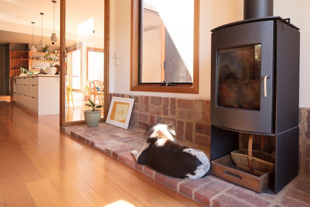 Warm, sunlit open-plan interior featuring a freestanding black wood-burning stove set on a raised red-brown brick hearth. A timber-framed window sits above the hearth, while the adjoining kitchen is visible in the background with white cabinetry, open timber shelving, pendant lights and timber flooring. A dog is resting on the hearth.