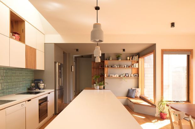 A warm, contemporary open-plan kitchen and dining area. The kitchen features a long white island and white cabinetry, a green textured tile splashback, timber open shelving, and integrated appliances. The adjoining dining area has timber-framed windows, a built-in window seat, a timber dining table, and light wood chairs. Soft pendant lights, plants, and shelving create a welcoming, sunlit interior.