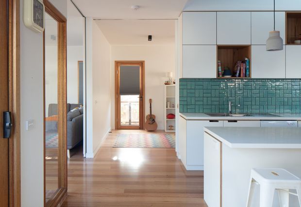 A bright open-plan kitchen viewed from the entry area, with white flat-panel cabinetry, a large island, and a distinctive teal-green geometric tiled splashback. Warm timber flooring and timber-framed openings lead toward a hallway and a partially visible living or bedroom area. Pendant lighting, open shelving, and a guitar add casual residential character.