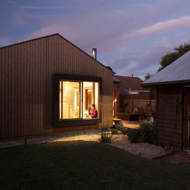 Dusk view of a compact timber-clad residential extension or outbuilding with a simple gabled roof and large warmly lit window. The building sits within a landscaped courtyard or garden, with gravel edging, planted beds, paving, outdoor seating and adjacent older structures visible. A person is seated at a desk or table inside, seen through the window.