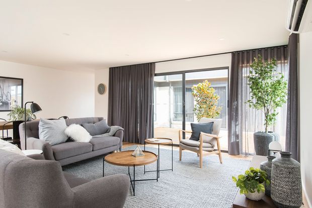 A bright, contemporary living room with a grey upholstered sofa and armchair, layered round timber coffee tables, a textured grey area rug, and a light timber lounge chair. Full-height dark grey curtains frame large glazed doors or windows opening to a planted courtyard. Neutral walls, indoor plants, soft furnishings, and restrained decor create a calm, refined atmosphere.