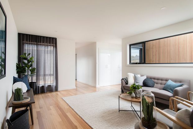 Bright contemporary living room with pale timber flooring, a large neutral textured rug, grey tufted sofa, black-framed coffee table, and light upholstered armchairs. Full-height sheer curtains and a high horizontal window with timber screening provide filtered natural light. Minimal white walls and clean-lined built-in elements create an airy, refined interior.
