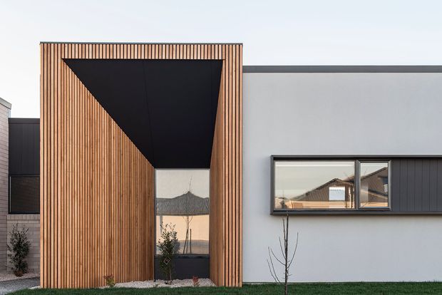 Front exterior view of a contemporary residential building with a sculptural entry volume clad in vertical timber battens. The entry is framed by a deep black, angular recessed canopy and tall glazed opening, alongside smooth light-grey rendered walls and dark horizontal window framing. A dark vertically clad wing and sparse landscaping complete the minimalist facade.