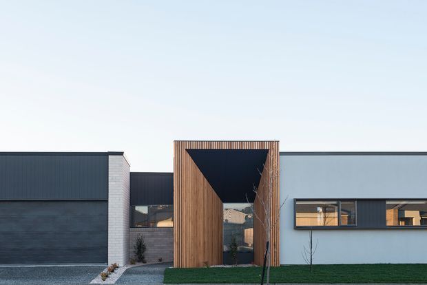 Contemporary single-storey residential facade with a strong geometric composition. Dark vertically ribbed cladding and a charcoal garage-like volume sit beside a pale rendered wall, while a central timber-clad portal frames a recessed entry and glazed opening. A narrow lawn, gravel path, and sparse planting complete the minimalist frontage.
