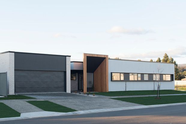 Street-facing view of a low, contemporary single-storey building with a flat roof and strongly composed geometric facade. The exterior combines pale masonry at the left, dark vertical cladding and a large dark garage door, a recessed entry framed in warm timber, and a light grey rendered facade with a horizontal band of windows. A gravel forecourt, manicured lawn strips, sidewalk, and road are visible in front.