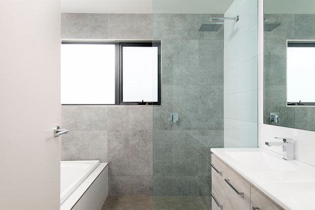 A clean, contemporary bathroom with large-format grey stone-look wall and floor tiles, a white bathtub, frameless glass shower enclosure, wall-mounted shower fixtures, and a white vanity with light timber cabinetry and chrome tapware. A black-framed window and large mirror add contrast and brightness.