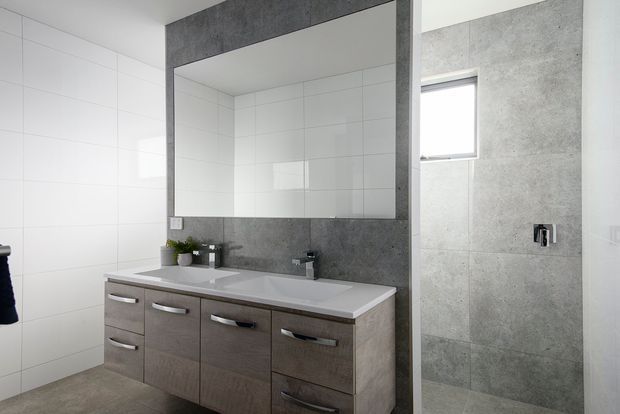 Contemporary bathroom featuring a wide floating-style vanity with grey wood-look cabinetry, a white integrated benchtop, large frameless mirror, white wall tiles, and a separate walk-in shower lined with grey concrete-look tiles. The room is bright and clean with simple modern fittings and a small high window.