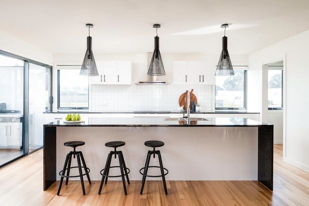 Bright, spacious contemporary kitchen with a large central island, three black bar stools, white flat-panel cabinetry, white tiled splashback, dark stone island ends and benchtop, timber flooring, and three black pendant lights. Extensive glazing and an open connection to adjacent areas create an airy, refined interior.