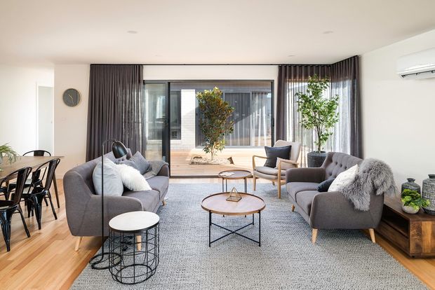 Bright, contemporary open-plan living and dining area with grey upholstered sofas and armchairs arranged around nested coffee tables on a large textured rug. A timber dining table with black chairs sits to the left, while full-height dark curtains frame black-framed glazing and a small courtyard beyond. Light timber flooring, pale walls, indoor plants, and a wall-mounted air conditioner complete the space.