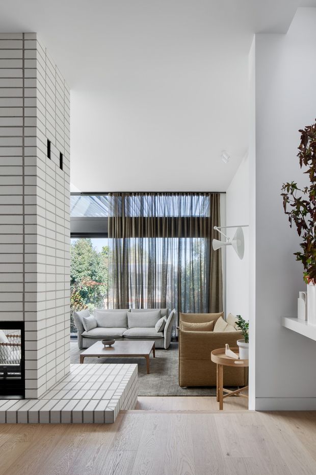 Bright, refined contemporary living room with a double-height white interior and extensive glazing overlooking greenery. A white tiled fireplace volume and hearth define the left side, while a pale timber floor, grey sofa, tan armchair, low timber coffee table, sheer dark curtains, and built-in white shelving create a calm, minimalist setting. A sculptural wall light and indoor tree add restrained decorative interest.