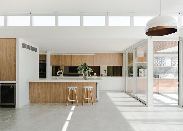 Bright contemporary open-plan kitchen with light timber cabinetry, a white island, integrated appliances, and two timber bar stools. Full-height glazed sliding doors open to an outdoor area, while high clerestory windows and a large sculptural pendant bring abundant daylight into the space. The polished concrete floor and restrained neutral palette create a clean, airy interior.