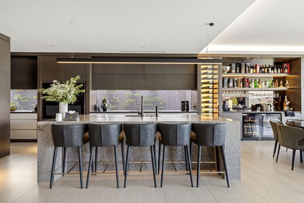 A spacious contemporary open-plan kitchen and entertaining area with a large stone island bench and extensive seating for eight. Dark timber cabinetry, integrated appliances, a pale stone benchtop, and a matching splashback create a refined, high-contrast fit-out. A fully stocked built-in bar and wine display sit to the right, while dining furniture is visible beyond the kitchen.