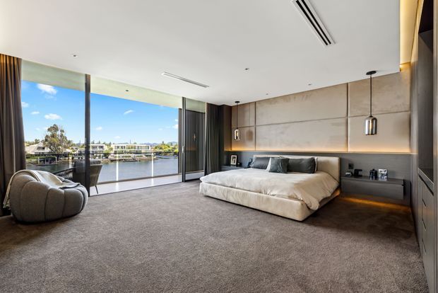 Spacious contemporary bedroom with floor-to-ceiling glazing overlooking a waterfront setting. The room features a large upholstered platform bed, sculptural lounge chair, dark built-in cabinetry, a full-width textured feature wall with integrated bedside shelving and warm concealed lighting, pendant lights, and a soft grey carpet. The restrained palette and expansive glazing create a refined, calm atmosphere.