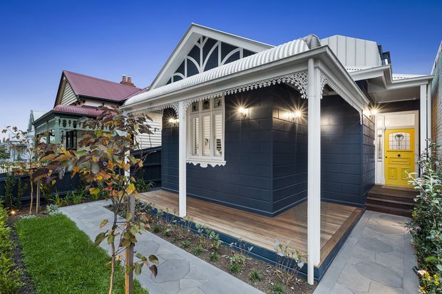 Exterior view of a renovated or newly built residential cottage-style home, featuring dark blue horizontal cladding, white decorative timber trim, a gabled front section with ornate fretwork, a corrugated metal roof, covered verandah, timber decking, stone-paved paths, and a bright yellow front door. A neighbouring traditional cottage is visible to the left.