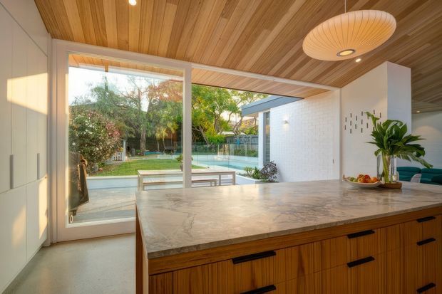 Warm contemporary kitchen with a large marble-look island, vertical timber cabinetry, white walls, and a timber-lined ceiling. Floor-to-ceiling glazing opens toward a landscaped garden and swimming pool, while a sculptural pendant light and indoor plants add visual interest.