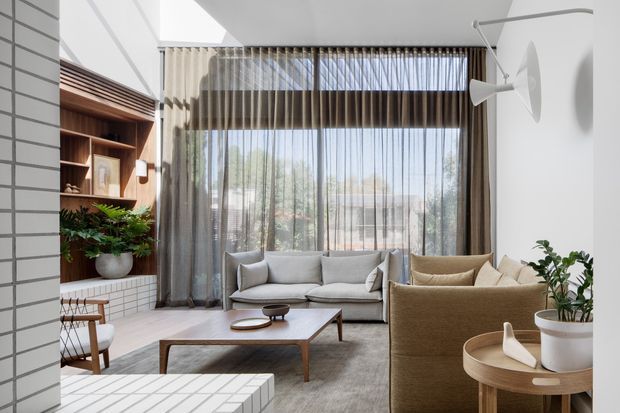 Bright contemporary living room with a light grey sofa, textured tan armchairs, a low timber coffee table, and large floor-to-ceiling glazed doors screened by sheer curtains. Built-in dark timber shelving and cabinetry line the left wall above white brick or tiled surfaces, while a white tiled fireplace or raised feature element occupies the foreground. The room has a high ceiling with strong natural light, indoor plants, and restrained mid-century-influenced furniture.