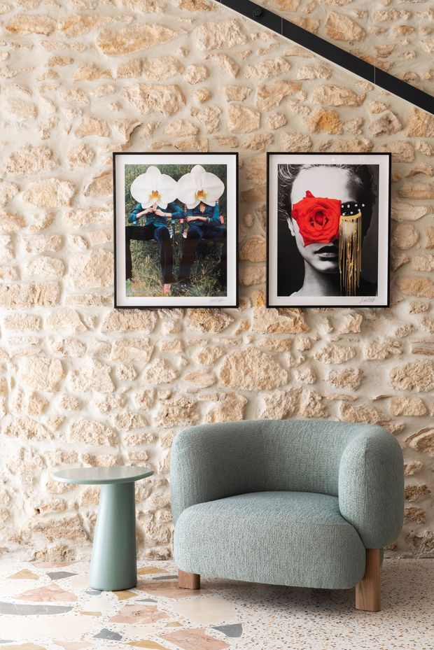 A styled seating vignette featuring a pale sage-green upholstered lounge chair and a small matching side table against a rustic exposed pale stone wall. Two framed artworks hang above the chair, and the floor is finished in multicoloured terrazzo. A black diagonal architectural element crosses the upper right corner.