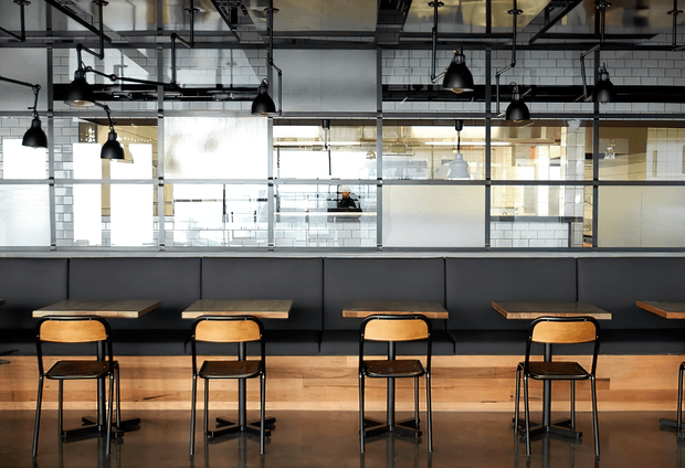 Industrial-style commercial interior with a row of small timber tables and black metal chairs arranged along a long dark upholstered banquette. The space features exposed ceiling services, suspended black pendant lights, black-framed glazed partitions, and white brick walls, creating a contemporary café, hospitality, or collaborative workplace setting.