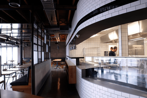 Interior view of a modern industrial-style restaurant or café, showing a curved white subway-tiled service counter with glass display panels, an open commercial kitchen, exposed dark ceiling structure and services, black-framed glazing, and timber-and-black dining furniture.