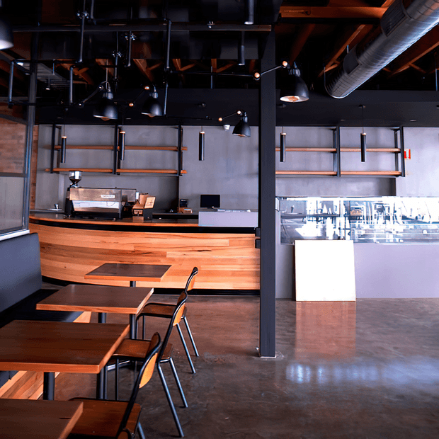 An industrial-style café or hospitality interior with a polished concrete floor, dark exposed ceiling structure, black steel columns and suspended lighting. A curved timber-clad service counter with espresso equipment occupies the rear, while timber tables and simple metal-framed chairs fill the foreground. A bright glazed display or bar counter is visible to the right.