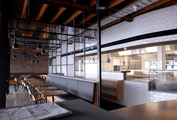 Industrial-style commercial dining space, likely a restaurant or café, with rows of timber tables and chairs, long upholstered banquette seating, exposed timber roof beams, black metal services and pendant lighting, and a bright tiled service counter and kitchen area separated by glazed partitions. The space combines dark structural elements with white subway tile and abundant daylight.