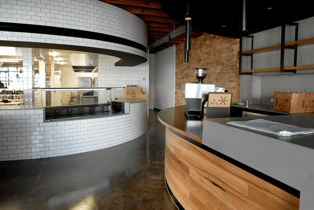 A contemporary commercial café or coffee shop interior with a curved white subway-tile service counter and open kitchen area. The foreground features a large curved timber-fronted counter with a dark benchtop, espresso equipment, shelving, exposed timber ceiling beams, a black ceiling, and a warm brick feature wall. The polished concrete floor and industrial food-service fit-out create a refined industrial atmosphere.