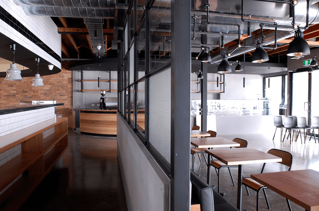 Industrial-style commercial interior with a café or restaurant counter on the left and an adjoining dining area on the right. The space features exposed services and ductwork, black metal-framed glazing, white subway-tile walls, exposed timber roof structure, brick accents, timber counters and tables, and black pendant lighting. Bright daylight enters through large windows at the rear.