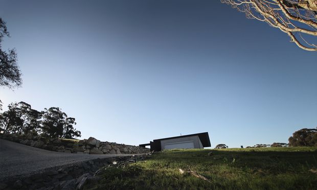 Low-angle exterior view of a minimalist single-storey building set into a gently sloping grassy landscape. The structure has a dark, sharply angular roof and dark exterior walls, with a large horizontal glazed opening facing the camera. A rough stone retaining wall and gravel driveway or path lead toward the building, while mature trees frame the scene against a clear blue sky.