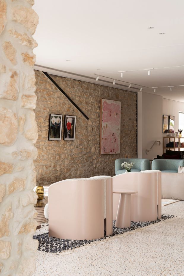 Bright contemporary living area with exposed rough stone walls, pale terrazzo flooring, and soft blush-pink and muted blue upholstered seating arranged around a small round side table. Track lighting and recessed ceiling lights illuminate artwork on the stone feature wall, while a stair or passage area is visible in the background. The space blends historic masonry with refined modern furnishings and a restrained pastel palette.