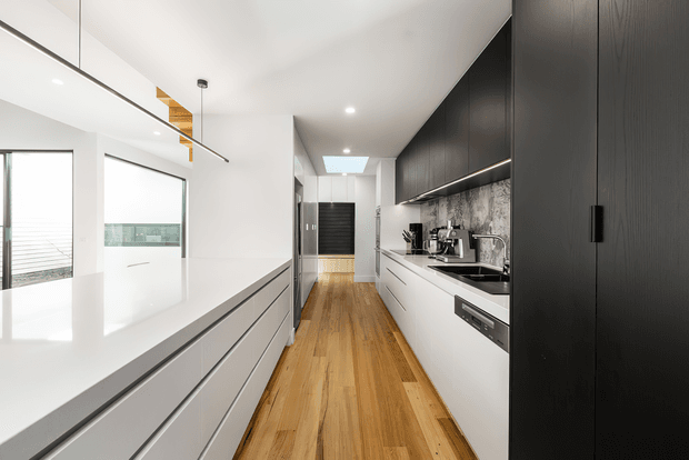 A bright, narrow contemporary galley kitchen with extensive white and matte black fixed cabinetry, a long white benchtop, dark patterned backsplash, integrated appliances, and warm timber flooring. A large glazed opening and balcony railing are visible at the left, while linear and pendant lighting define the ceiling.