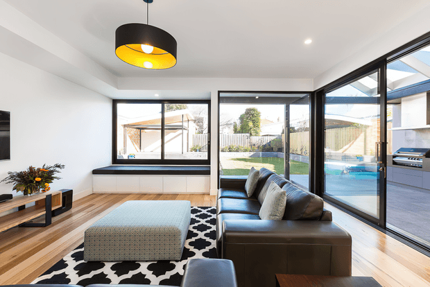 Contemporary open-plan living room with a large dark leather sectional sofa, patterned black-and-white rug, upholstered ottoman, timber flooring, and a low built-in window seat or storage unit. Full-height black-framed glazing opens views to a landscaped backyard and pool area, while a covered outdoor kitchen with a built-in grill is visible beyond. A black pendant light with a warm yellow interior provides a strong focal point.