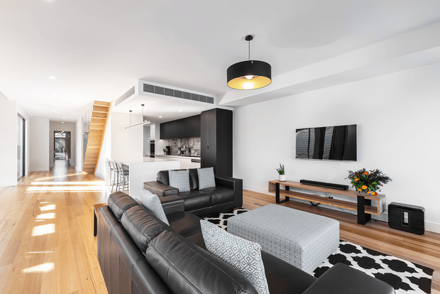 Bright, open-plan contemporary living room with polished timber flooring, black leather sectional seating, a large grey ottoman, patterned rug, wall-mounted television, and a timber media console. The room opens toward a compact kitchen with dark cabinetry, while a timber staircase and long hallway extend into the background. White walls and ceiling, recessed lighting, and a black pendant create a clean, modern interior.