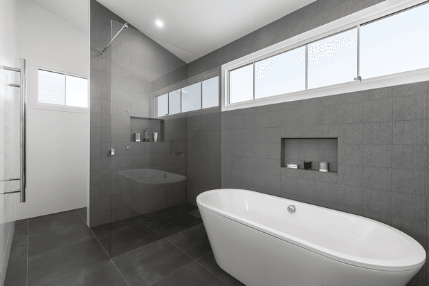 Contemporary bathroom with a freestanding white bathtub, frameless glass shower enclosure, wall-mounted vanity area, recessed wall niche, and extensive dark grey tiled wall and floor finishes. High horizontal windows provide abundant natural light, creating a clean, bright, minimalist atmosphere.