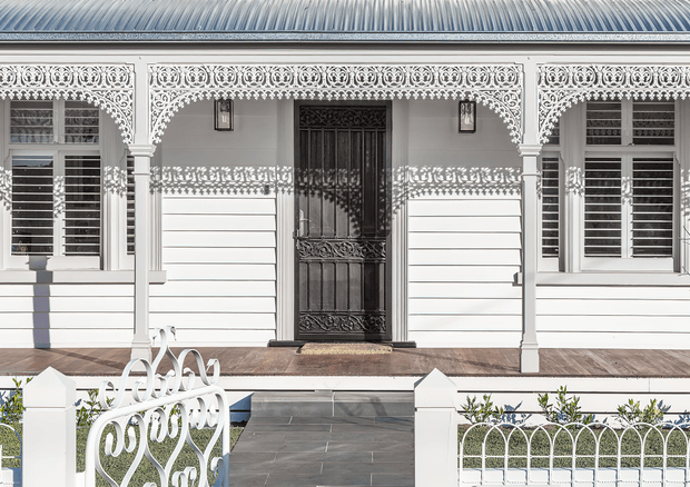 Front elevation of an ornate single-storey residential facade, featuring white painted weatherboard walls, elaborate decorative lacework veranda detailing, shuttered windows, a dark embellished entry door, timber porch decking, and a paved path leading to decorative white metal fencing and gates. The facade has strong heritage character and intricate period detailing.