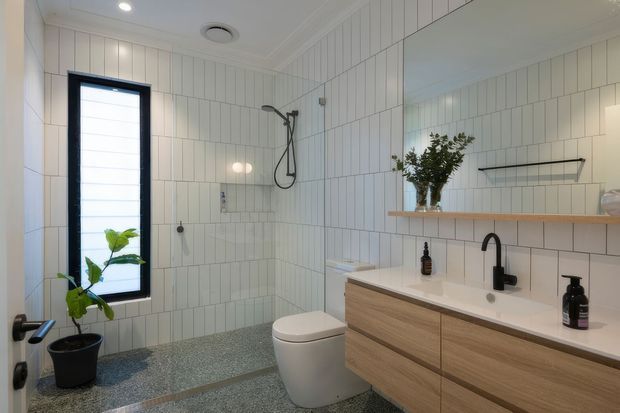 Bright contemporary bathroom with full-height white vertical wall tiles, a frameless walk-in shower, freestanding-style toilet, and a floating timber vanity with a white integrated basin. A large mirror, black tapware and shower fittings, open shelving, and a narrow black-framed window complete the space.