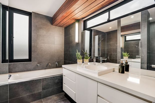 Contemporary bathroom with a freestanding-style bathtub, double vanity, large wall mirror, dark grey stone-look tiled walls and floor, and a warm timber-lined ceiling. Black-framed windows and glazing provide contrast, while white cabinetry and a white benchtop keep the room bright and refined.