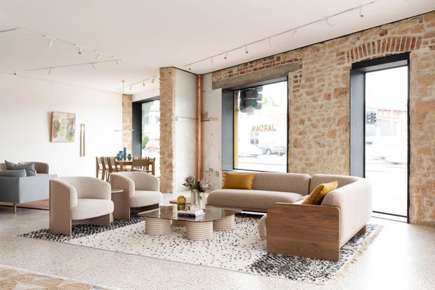 Bright open-plan interior with a lounge and dining area in a converted masonry building. Exposed irregular brick and stone walls, black-framed industrial windows and doors, white ceiling with track lighting, and warm timber accents surround contemporary upholstered seating, a low coffee table, and a compact dining setting.