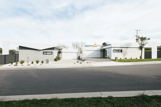 A street-facing view of a low-rise contemporary residential development consisting of two adjacent modern homes. The buildings feature crisp white rendered walls, dark contrasting roof planes and window frames, flat and shallow-pitched roof forms, broad concrete driveways, landscaped front setbacks, and tall dark privacy fencing. The image is photographed from across a paved residential street under a bright, lightly overcast sky.