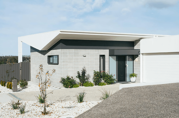 Single-storey contemporary home exterior with a low, angular flat roof, white projecting eaves, dark fascia, pale grey tiled facade, dark-framed glazing, and an integrated white garage. The front yard uses light gravel, white stone, and sparse drought-tolerant planting, with a broad paved driveway in the foreground.