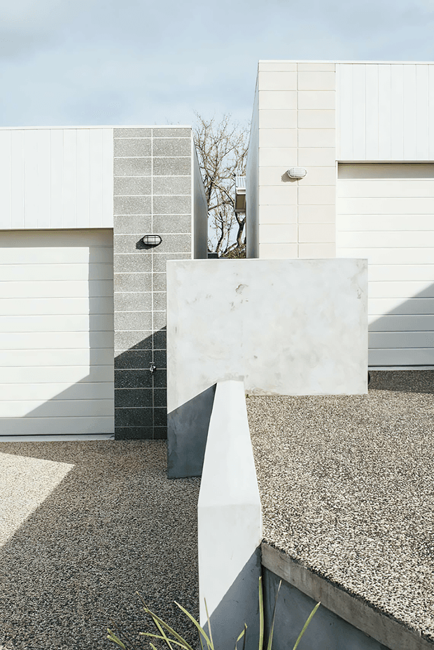 Exterior architectural photograph of a minimalist, geometric building composition. White horizontal-panel walls and garage-like doors are paired with a dark textured tiled vertical wall and a large pale concrete screen or sculptural privacy wall. The foreground includes a pebble-finished flat roof or terrace edge, strong sunlight, and sharp angular shadows. The image is dominated by clean rectilinear forms and a restrained white, grey, and charcoal palette.