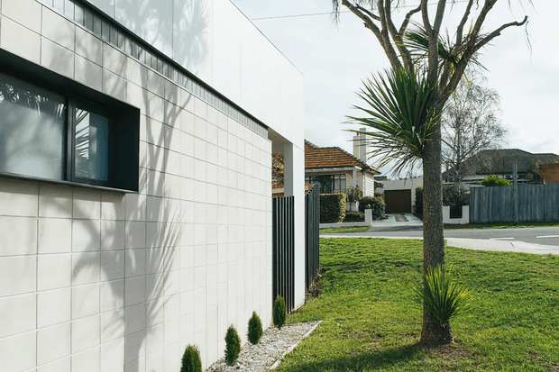 Exterior view of a contemporary residential building, featuring a clean white tiled or panelled facade, a dark-framed horizontal window, and a minimalist black vertical-slat side gate. The building sits within a suburban streetscape with a carefully maintained lawn, gravel planting strip, clipped shrubs, and sculptural palm-like planting. Traditional neighbouring homes and low fencing are visible beyond.