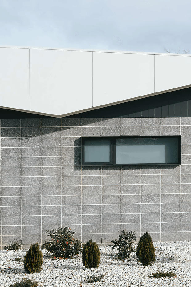Close-up exterior view of a modern building facade, featuring a dark grey textured rectangular tile wall, a projecting white parapet or roof volume, black-framed horizontal windows, and a minimalist gravel garden with clipped shrubs in the foreground.