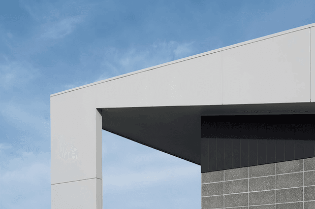 Minimalist modern architectural exterior detail showing a large white rectangular facade or portal with a deep dark soffit projecting over a grey blockwork wall. Clean horizontal and vertical panel joints create a stark geometric composition against a blue sky.