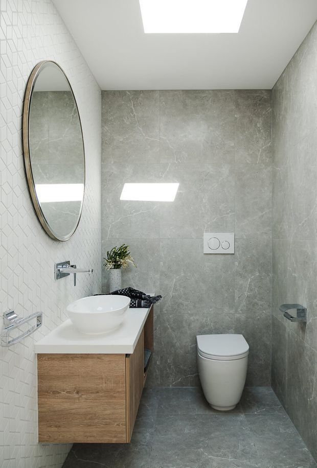 A compact contemporary bathroom with a wall-mounted timber vanity, white vessel basin, chrome tapware, large grey stone-look wall and floor tiles, patterned white feature tiles, a round framed mirror, wall-hung toilet and a rectangular skylight. The room is bright, clean and minimalist in appearance.