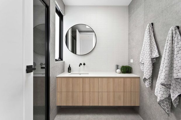 A contemporary bathroom viewed through an open door, featuring a floating timber-look vanity with integrated white benchtop, round wall mirror, wall-mounted black tapware, white rectangular wall tiles, grey textured wall finish, dark-framed shower glazing, and two patterned white towels. The room has a clean, refined, neutral palette with warm wood cabinetry.