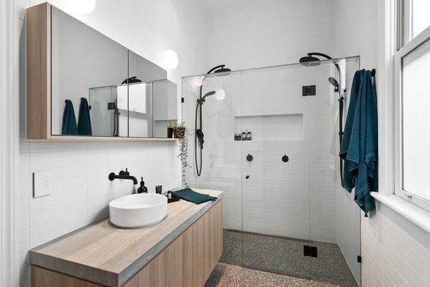 Bright contemporary bathroom featuring a walk-in glass shower with dual black fixtures, white wall tiles, a terrazzo-style shower floor, and a floating timber-look vanity with a round vessel basin. Large mirrors, black hardware, white walls, and a window create an airy, clean interior.
