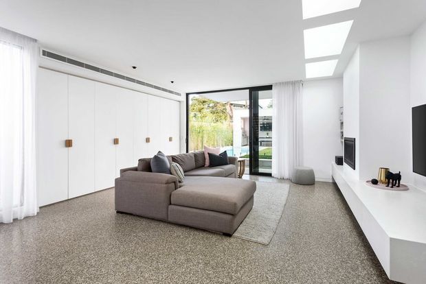 Bright contemporary living room with a large taupe sectional sofa and ottoman on a textured area rug. Full-height white built-in cabinetry with timber knobs lines the left wall, while a sleek white media console and wall-mounted television occupy the right. Floor-to-ceiling glazing with sheer curtains opens to an outdoor area, and several rectangular skylights bring additional daylight into the high, clean-lined space.